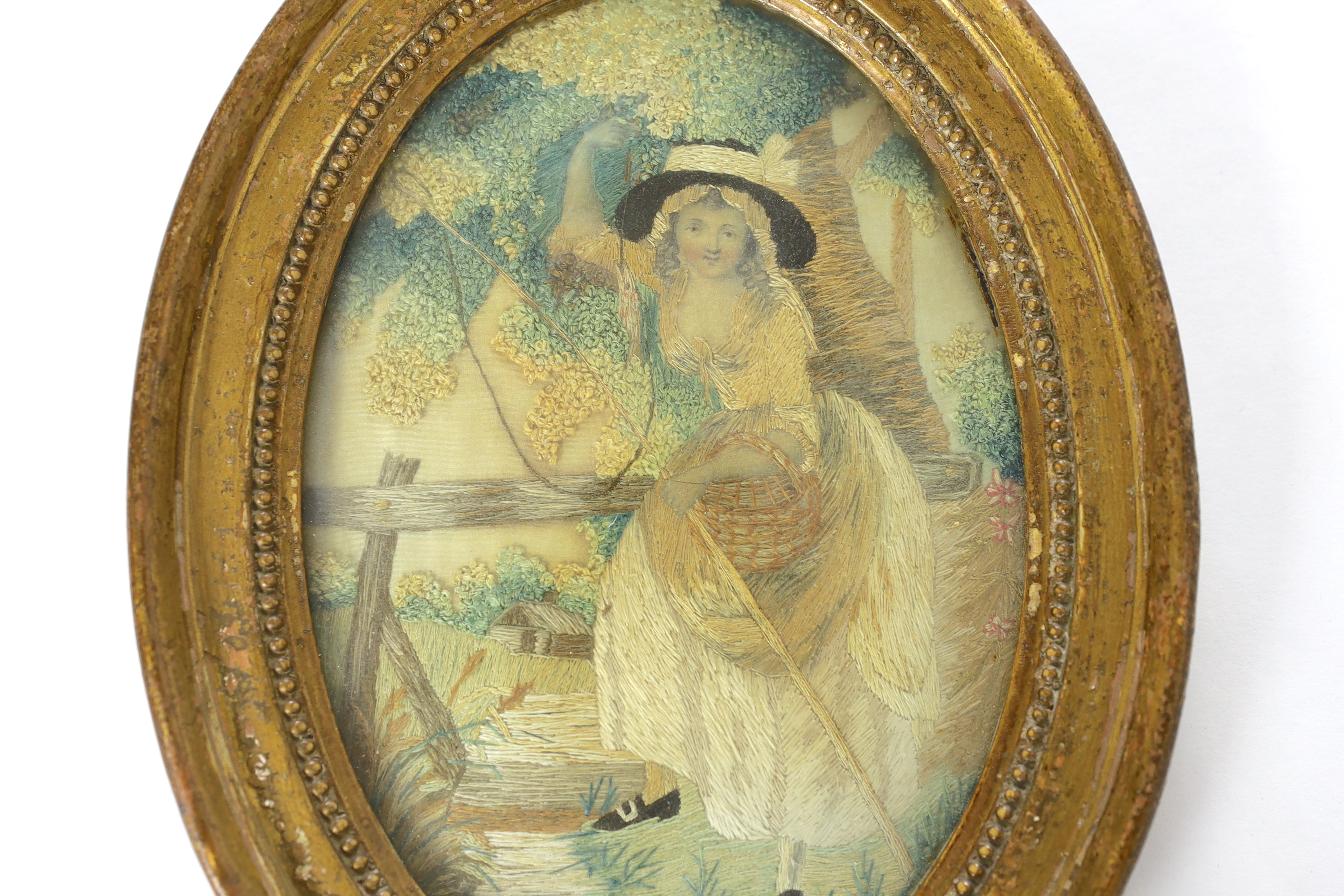 An early George IV oval silk work needlework pastoral embroidery, of a young girl with a fishing rod and basket under a tree by a bridge and stream, worked in fine wools and silks in stem stitch and a large knotting stit